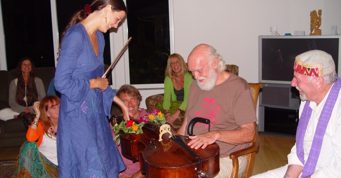 Ashana Sophia standing with a bow beside Ram Dass, who is seated with the flower-garlanded cello across his lap, in a living room of laughing friends