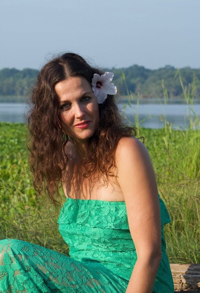 Ashana Sophia seated by a lake at golden hour in a green lace dress, a pale hibiscus in her hair, looking directly at the camera